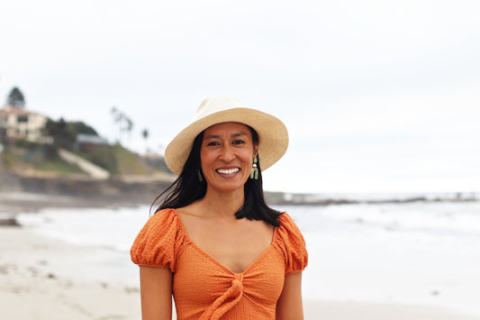 Photo of surf coach trained in surf therapy, Meg Aquinde in a tan sun hat wearing an orange short sleved blouse standing   on the beach in the san with ocean and cliffside backdrop